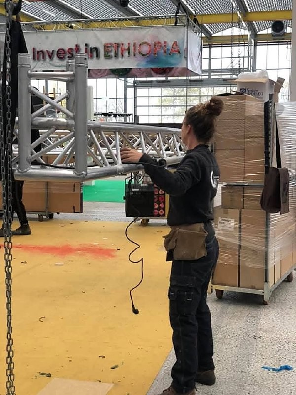 Vakvrouw positioneert een aluminium truss tijdens een opbouw op een beursvloer