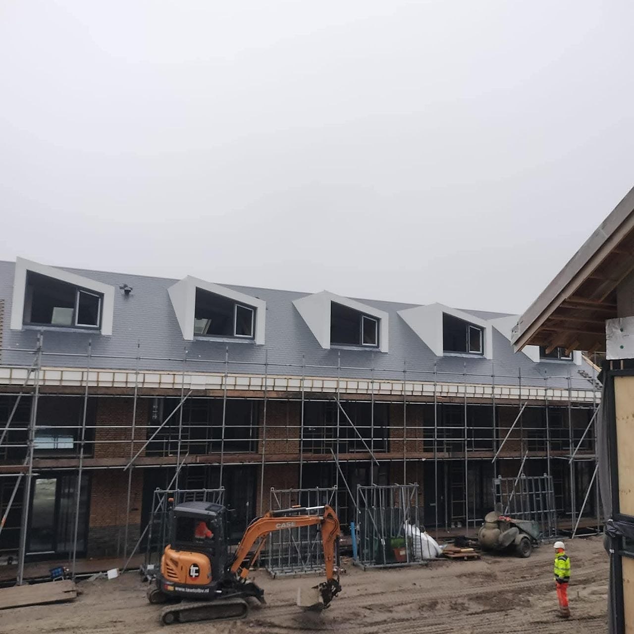 Rij woningen in aanbouw met witte dakkapellen, steigers en een minigraver op de bouwplaats