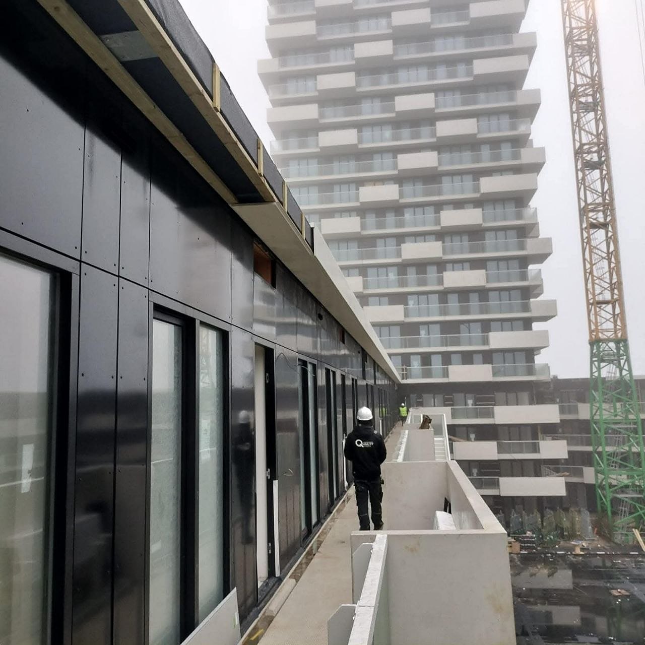 Quality Finish-medewerker op een galerij langs een zwarte gevel, met een woontoren en bouwkraan op de achtergrond