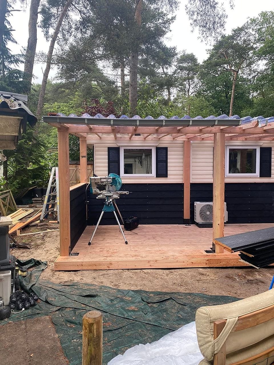 Houten overkapping met vlonder in aanbouw naast een chalet, met een afkortzaag op het terras