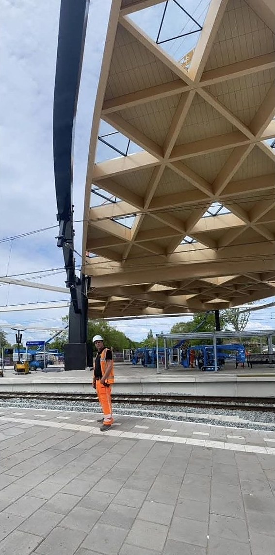 Houten dakconstructie met driehoekig vakwerk boven een perron, met een vakman in oranje werkkleding