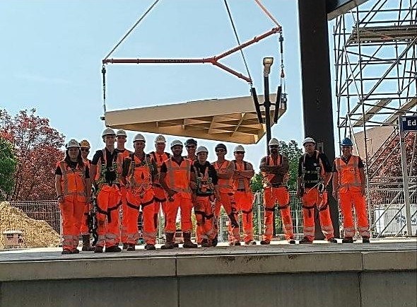 Team in oranje veiligheidskleding en helmen op een perron, met een hijsdeel van de dakconstructie aan een kraan